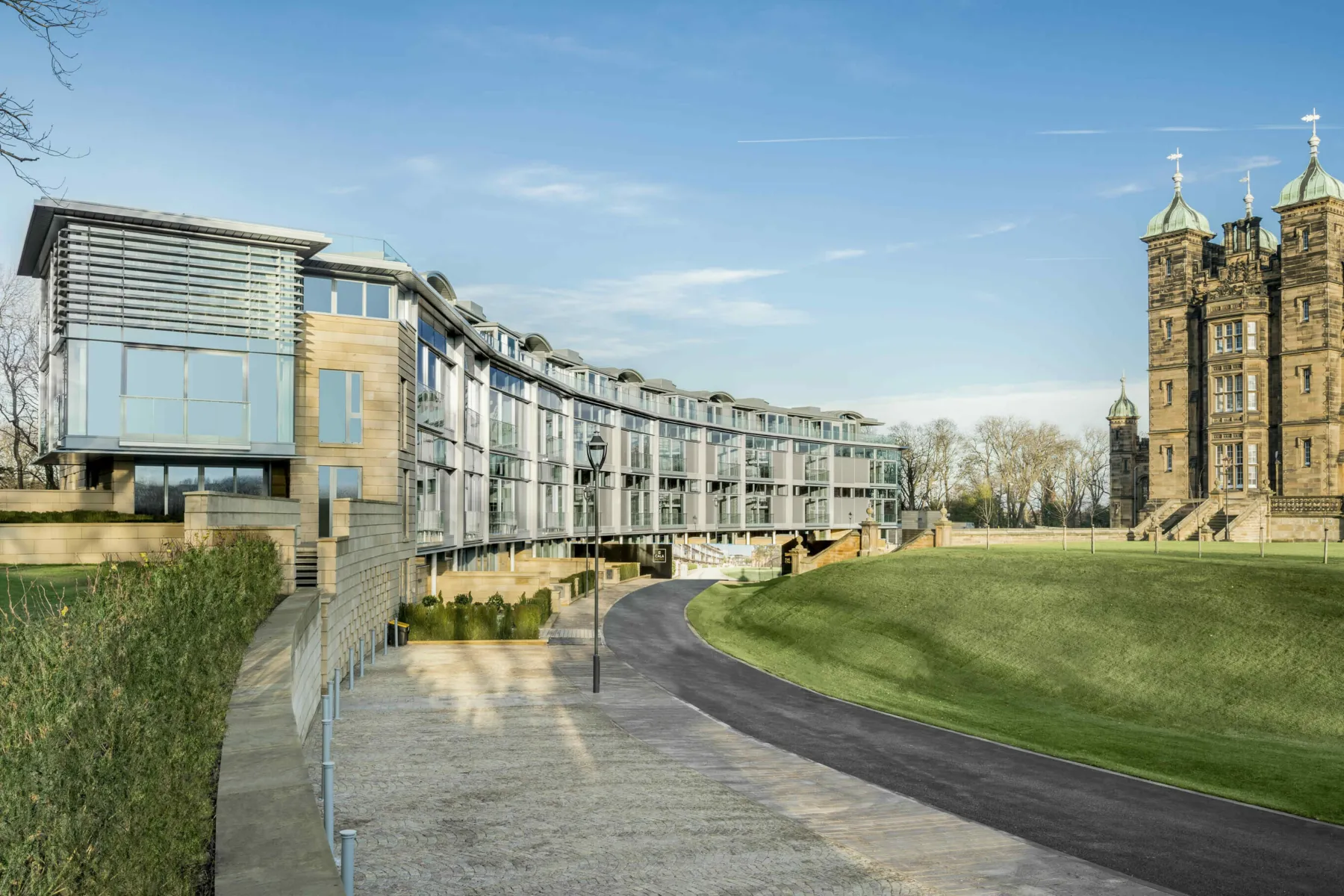 Exterior view of The Crescent at Donaldson's College, Edinburgh. A contemporary curve of apartments in glass and sandstone with three storeys plus penthouse. The building is set beside terraced lawns with corner towers of the original college building just seen