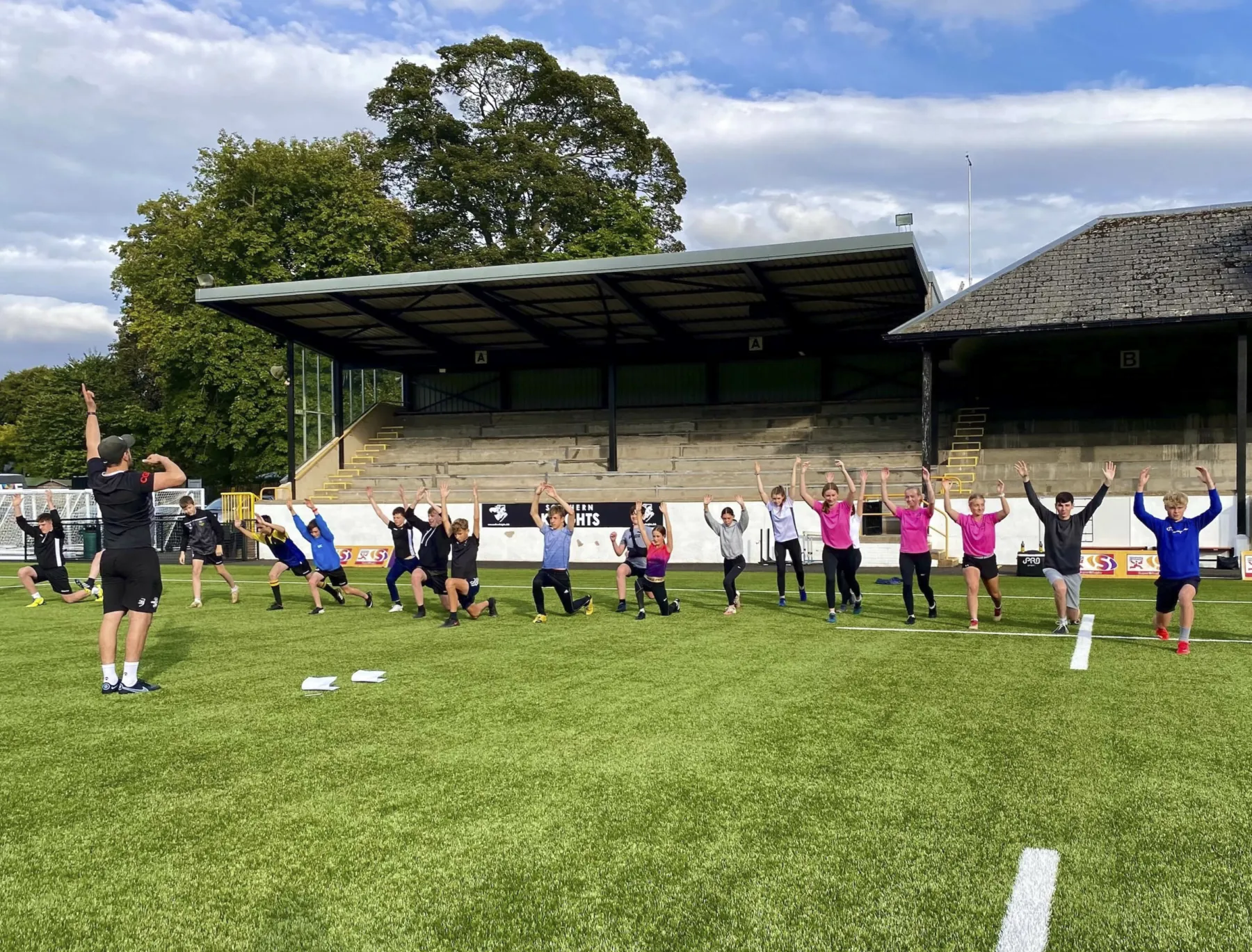 Melrose Athlete Development programme at the stadium in Melrose, Scottish Borders. A group of young people is led in a warm up by an instructor. It is a bright day in summer and trees are shown behind the stadium.