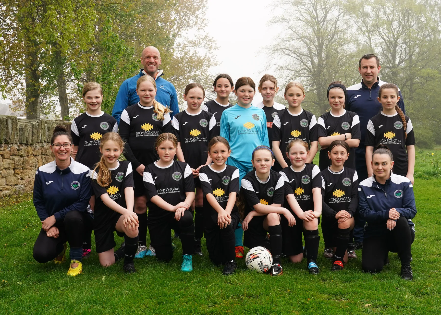 Dalkeith Thistle football team for girls under 12. The team lines up with its coaches and sponsor for a team photo wearing their black strip.