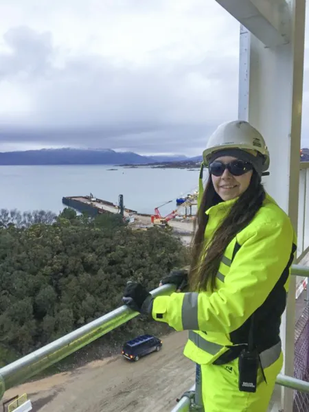 A young woman in hi-vis clothing and hard hat, working as a construction consultant, on one of the upper levels of a building under construction on the Isle of Skye, Scotland. Behind her is a view to the pier at Kyleakin, sea and hills.