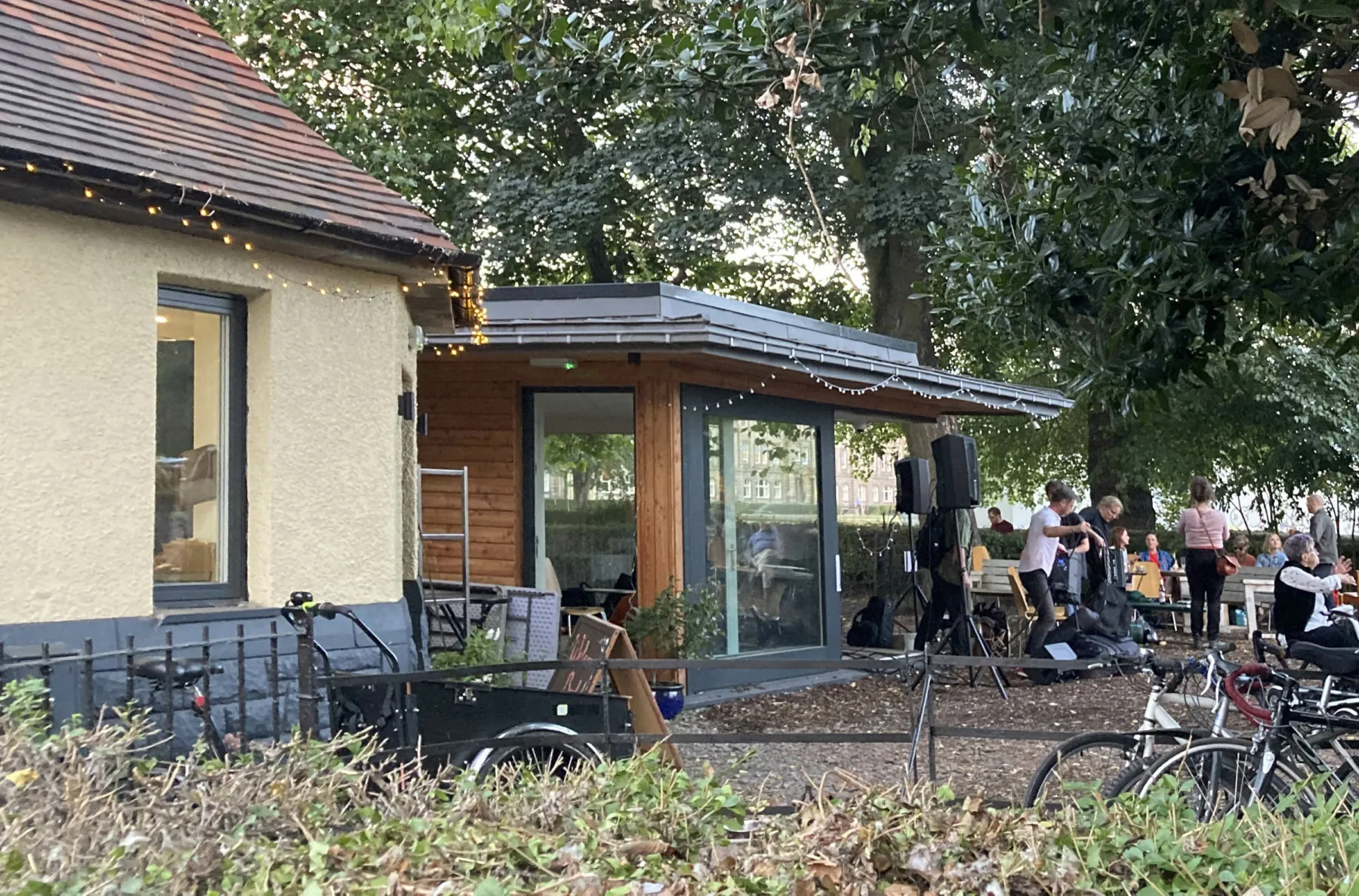 View of the new extension at Leith Community Croft, an urban growing project with cafe. The new extension is glazed and has a flat sedum roof. Visitors gather under the trees.