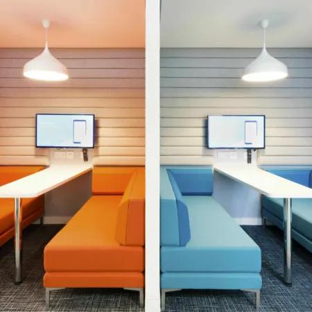 Interior at 102 Westport, offices in Edinburgh. Two booths with banquette seating - one in orange and one in blue. A central white table has a screen at the end in each, and white pendant lamps sit above the tables.