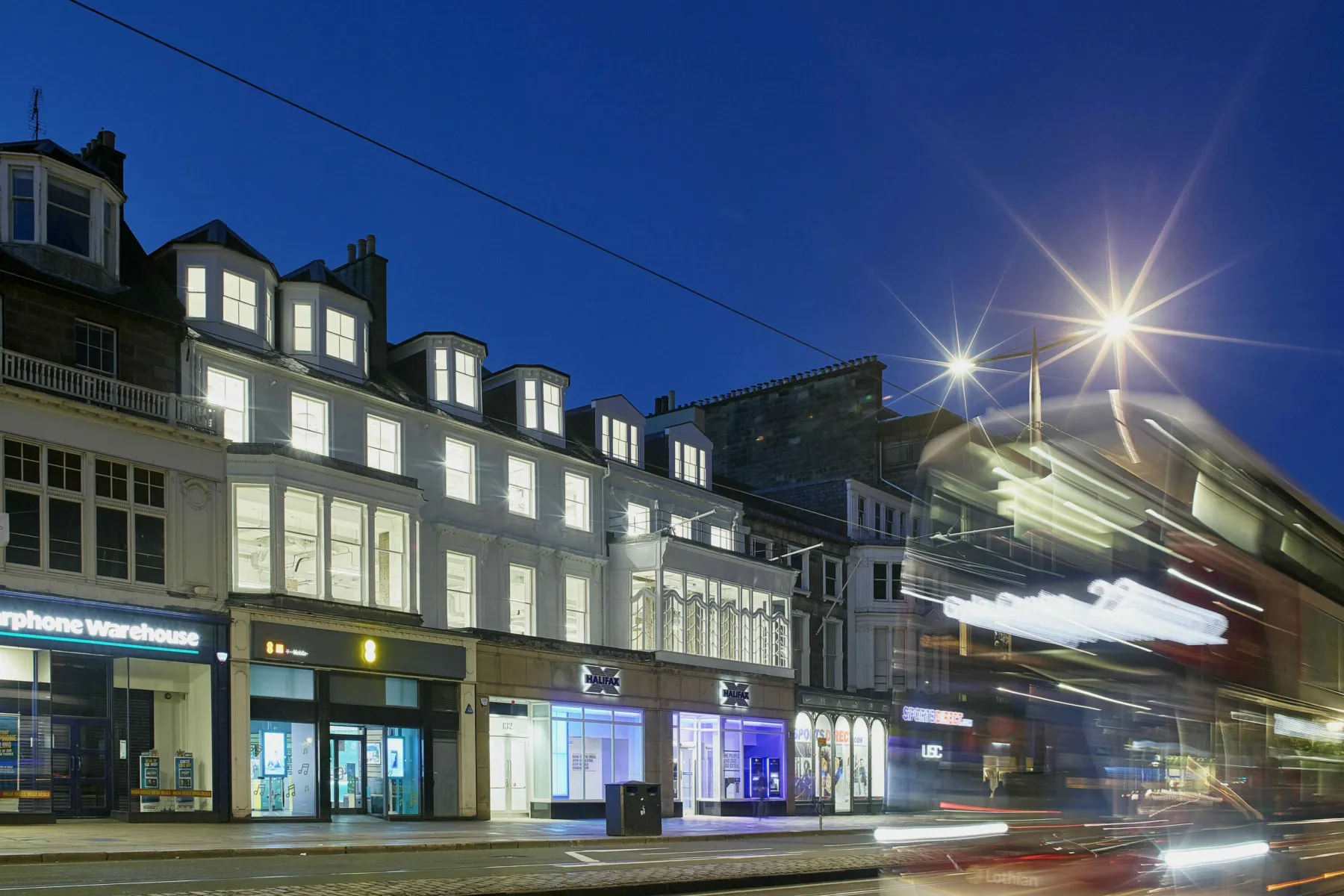 A nighttime view of Princes Street, Edinburgh. In the foreground a bus moves (shown out of focus, in motion). In the mid ground a range of offices including the listed building at 132 Princes St. The upper three storeys are refurbished office accommodation and are lit up from within.