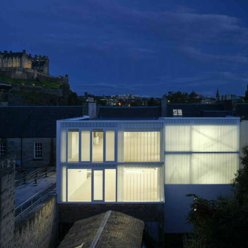 A modern, glass box office constructed in two sections behind a listed building in the city centre of Edinburgh. Shown at night, with the buildings glowing and with Edinburgh Castle in the background. The building is empty of furniture.