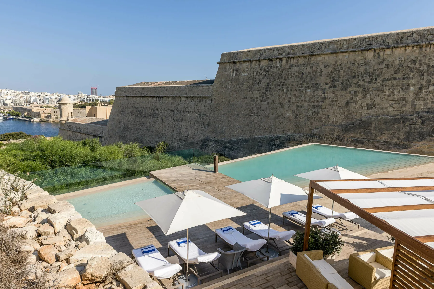 The outdoor pool and terrace at Hotel Phoenicia, Malta. Bordered by a historic city wall, the terrace has views across Valletta and a garden. Loungers and parasols are arranged by the pool.