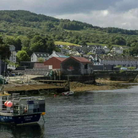 The pier and Egg Shed at Ardrishaig. In the foreground a small fishing boat, behind it the town and the Egg Shed by the shore - clearly visible with its rusty orange roof and cladding.
