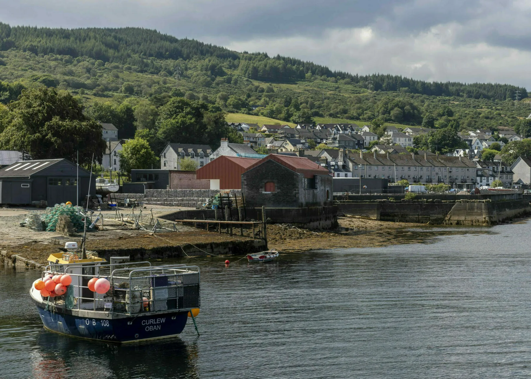 The pier and Egg Shed at Ardrishaig. In the foreground a small fishing boat, behind it the town and the Egg Shed by the shore - clearly visible with its rusty orange roof and cladding.