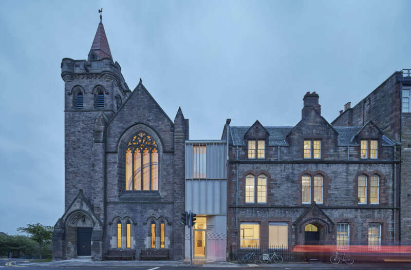 Exterior view of the Greyfriars Charteris Centre after extensive conservation work to the historic church building, refurbishment and extension. Between the former church and sandstone offices there is now a contemporary extension in timber with terrazzo render. the photograph shows twighlight with traffic and people passing - blurred with motion.