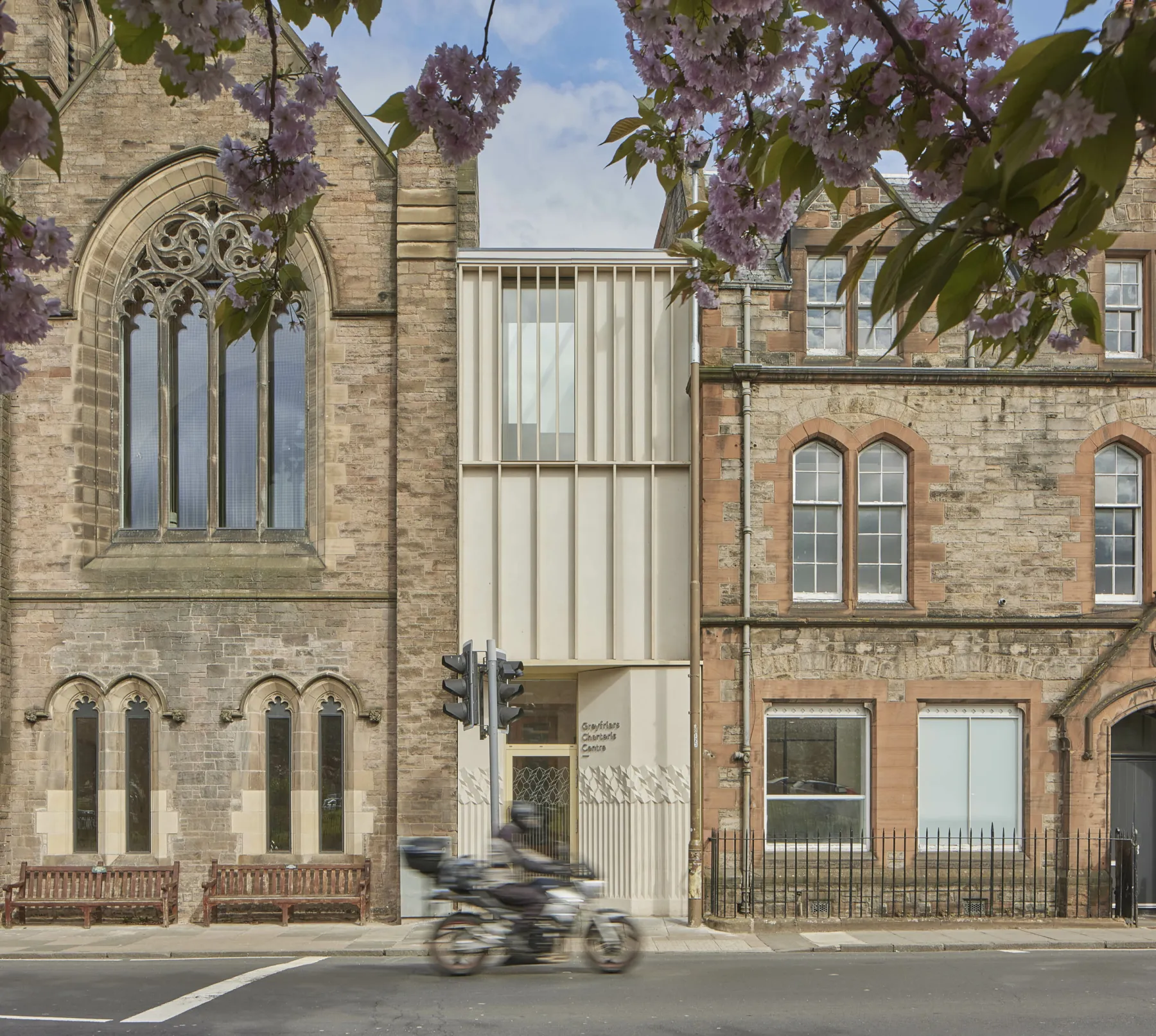 Exterior of the restored and extended buildings at Greyfriars Charteris Centre Edinburgh. A motorcyclist passes the door to the new extension, which is timber clad in terrazzo. The restored church is on the left, with the victorian three storey sandstone office building on the right. Cherry blossom frames the image.