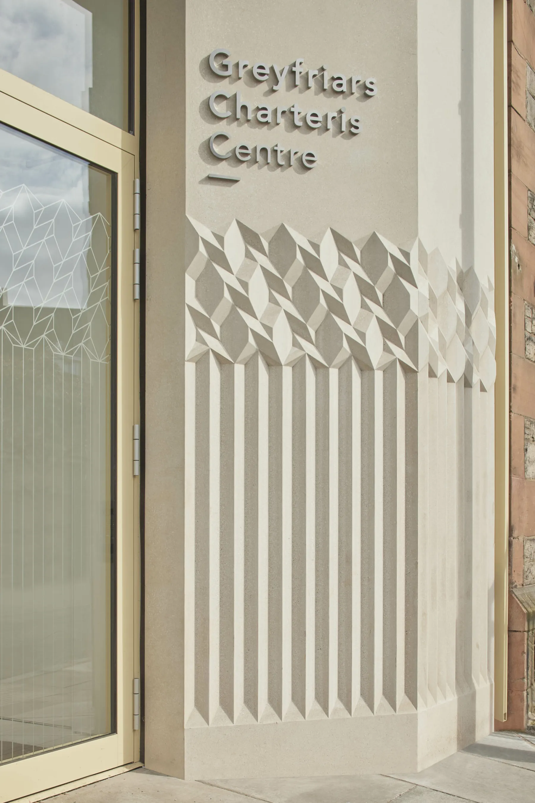 Detail of the pale, sandy coloured terrazzo cladding at Greyfriars Charteris Centre, Edinburgh. Textured decoration sits beneath the sign. To the left, the glass door at the entrance.