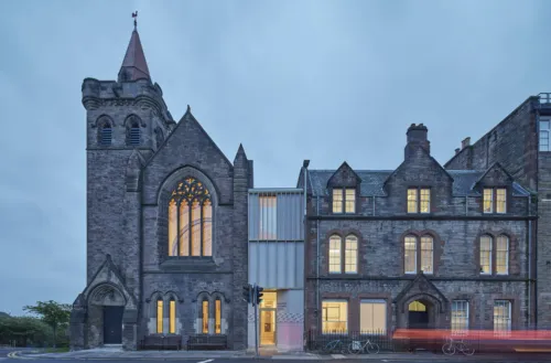 Exterior view of the Greyfriars Charteris Centre after extensive conservation work to the historic church building, refurbishment and extension. Between the former church and sandstone offices there is now a contemporary extension in timber with terrazzo render. the photograph shows twighlight with traffic and people passing - blurred with motion.