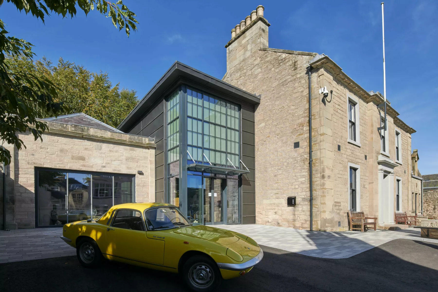 Exterior view of the Jim Clark Motorsport Museum at Duns in the Scottish Borders. The museum is made up of several historic buildings including a sandstone, Georgian detached house and several outbuildings. The garage has been linked to the house with a contemporary structure clad in dark grey. Outside a yellow, Lotus Elan S3 Coupe vintage sports car is parked in the driveway.