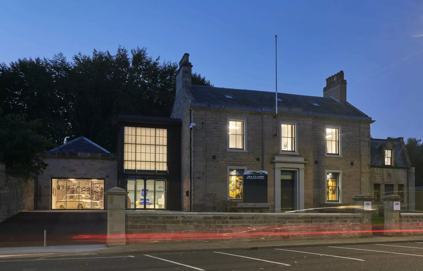 Exterior, nighttime view of the Jim Clark Motorsport Museum, Duns, Scottish Borders. The building is lit up within - a sandstone Georgian villa with outbuildings now linked via a contemporary, double height construction. Tail lights trail across the image, a car has passed by.