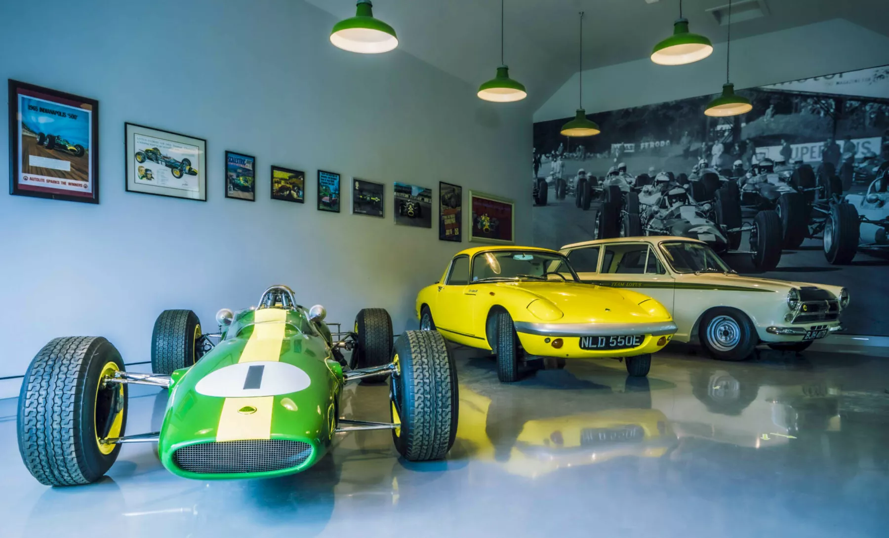 Inside the garage at the Jim Clark Motorsport Museum showing vintage Lotus sports cars all driven by Jim Clark (a green formula one Lotus Type 25, a yellow Lotus Elan S3 Coupe, a cream Lotus Cortina) and wall displays of historic motor racing photographs.