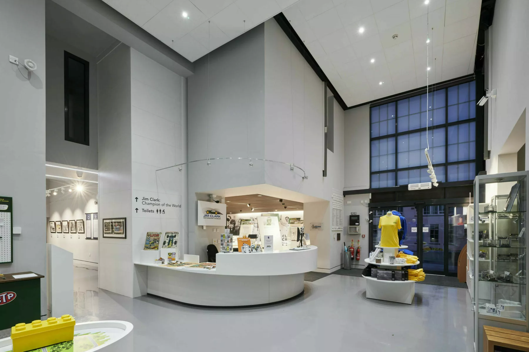 Inside the entrance atrium at the Jim Clark Motorsport Museum, Scottish Borders. A double height space with curved reception desk and displays of merchandise. Through a doorway is one of the exhibition rooms.