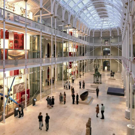 Interior of the main hall at the National Museum of Scotland, Edinburgh after reburbishment. The hall is a victorian iron construction, with three storeys of galleries around an atrium. The roof is glass. Visitors mill around on the ground floor and the other levels. Some exhibits are back-lit.