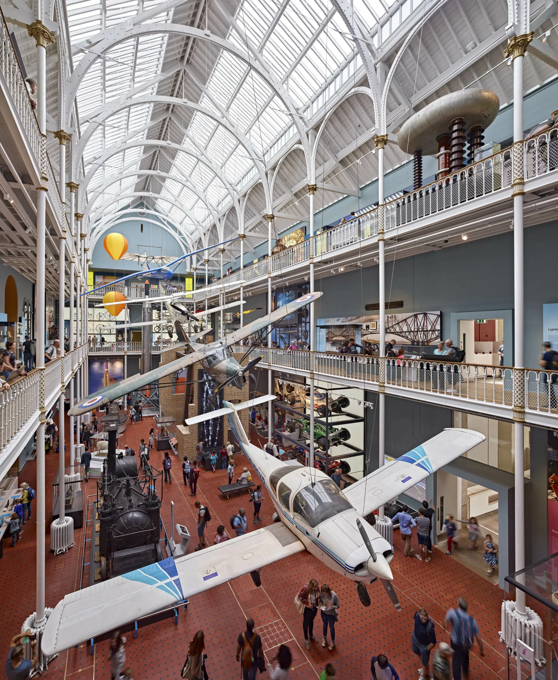 Inside the National Museum of Scotland Science and Technology Galleries on Chamber Street, Edinburgh. A large victorian hall with three storeys, galleries around the main atrium, wrought iron pillars and a glazed ceiling. Small aircraft hang into the space, suspended from the rafters. Visitors mill around all the spaces.