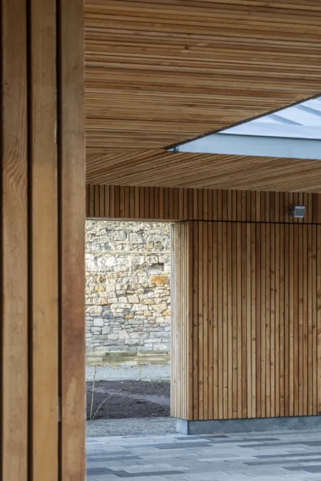 A view through a contemporary, timber lined shelter towards an old stone wall. The building has a skylight and a stone floor. At Saughton Park and Gardens, Edinburgh,