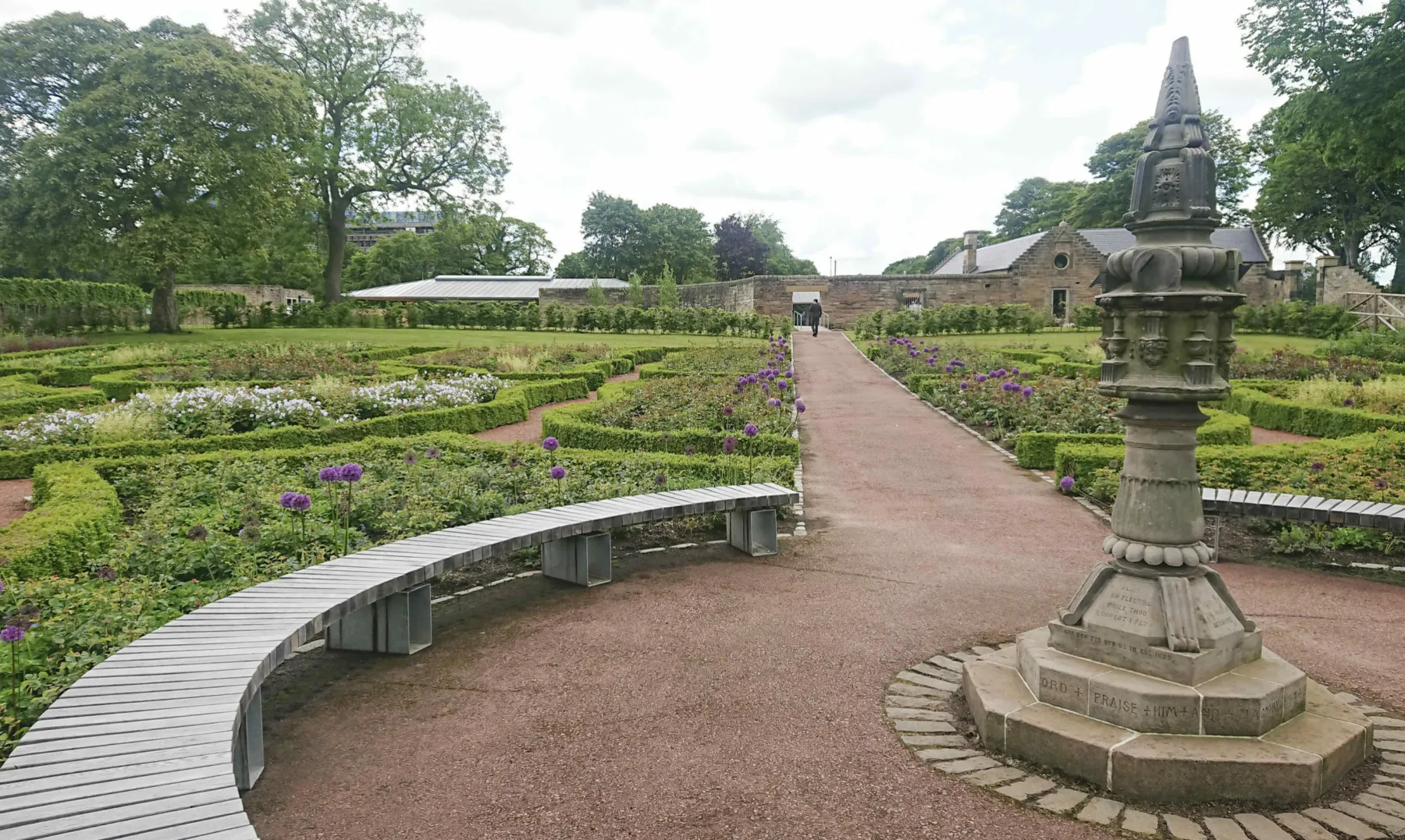 Saughton Park and Gardens, Edinburgh. In the formal garden a historic sandstone sundial is surrounded by  a ring of low wooden benches and flower beds edged with box hedging. In the distance the buildings of Saughton Park - cafe and visitor centre.