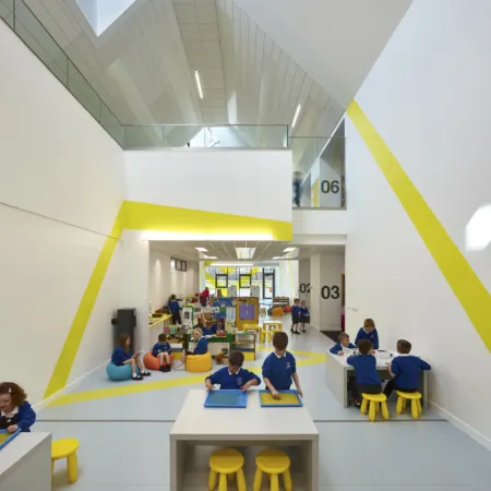 Inside Broomlands Primary School in a double height area with raised crossing. Children sit at tables for activities and in a storytelling area.