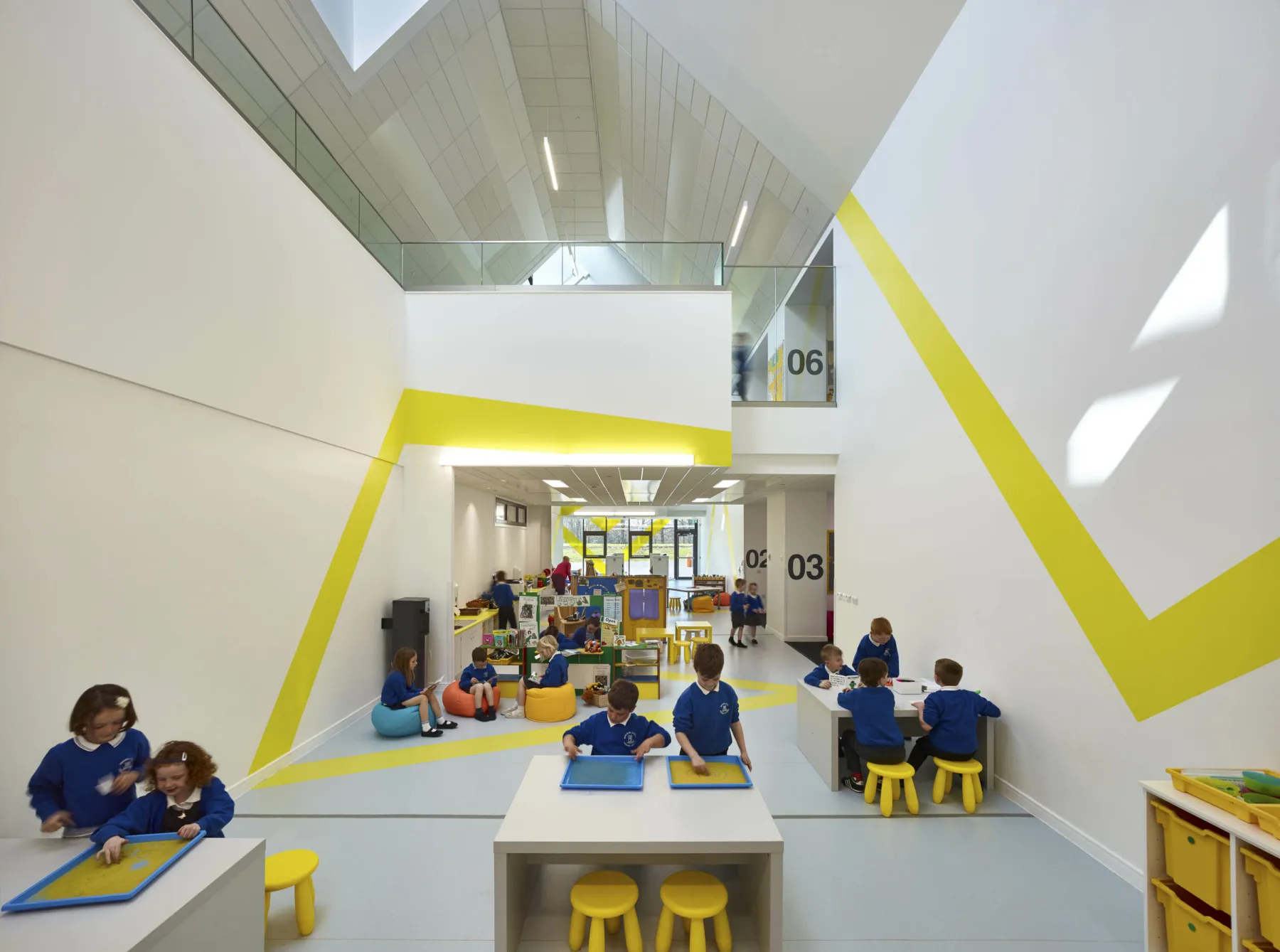 Inside Broomlands Primary School in a double height area with raised crossing. Children sit at tables for activities and in a storytelling area.
