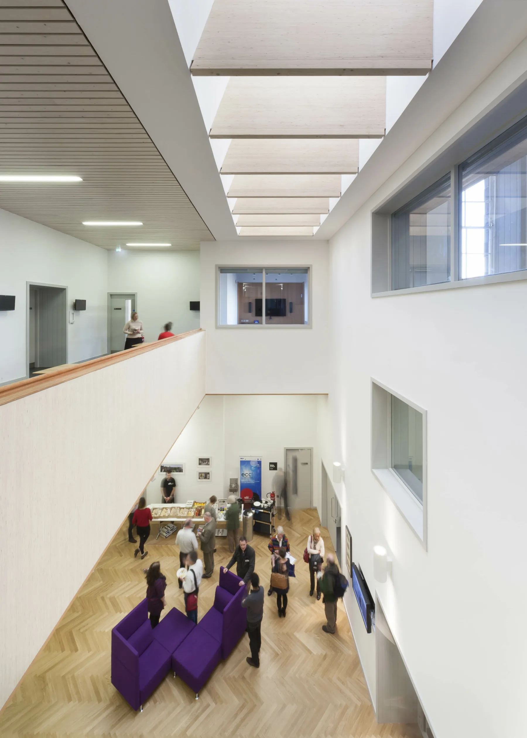 Looking down into the central atrium at the Edinburgh Climate Change Institute from the first floor gallery. A group takes part in a reception with refreshments. The floors are herringbone timber, walls are white and ceiling and skylights have timber detailing. It is a brightly lit space.