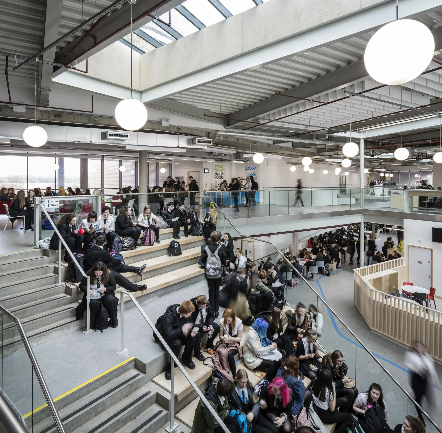In the central atrium at Garnock School, Scotland. Pupils sit beside the stairs on bench seating, and queue in the cafeteria. The design features exposed services, concrete and large skylights. One of the projects of Thomson Gray Construction Consultants.