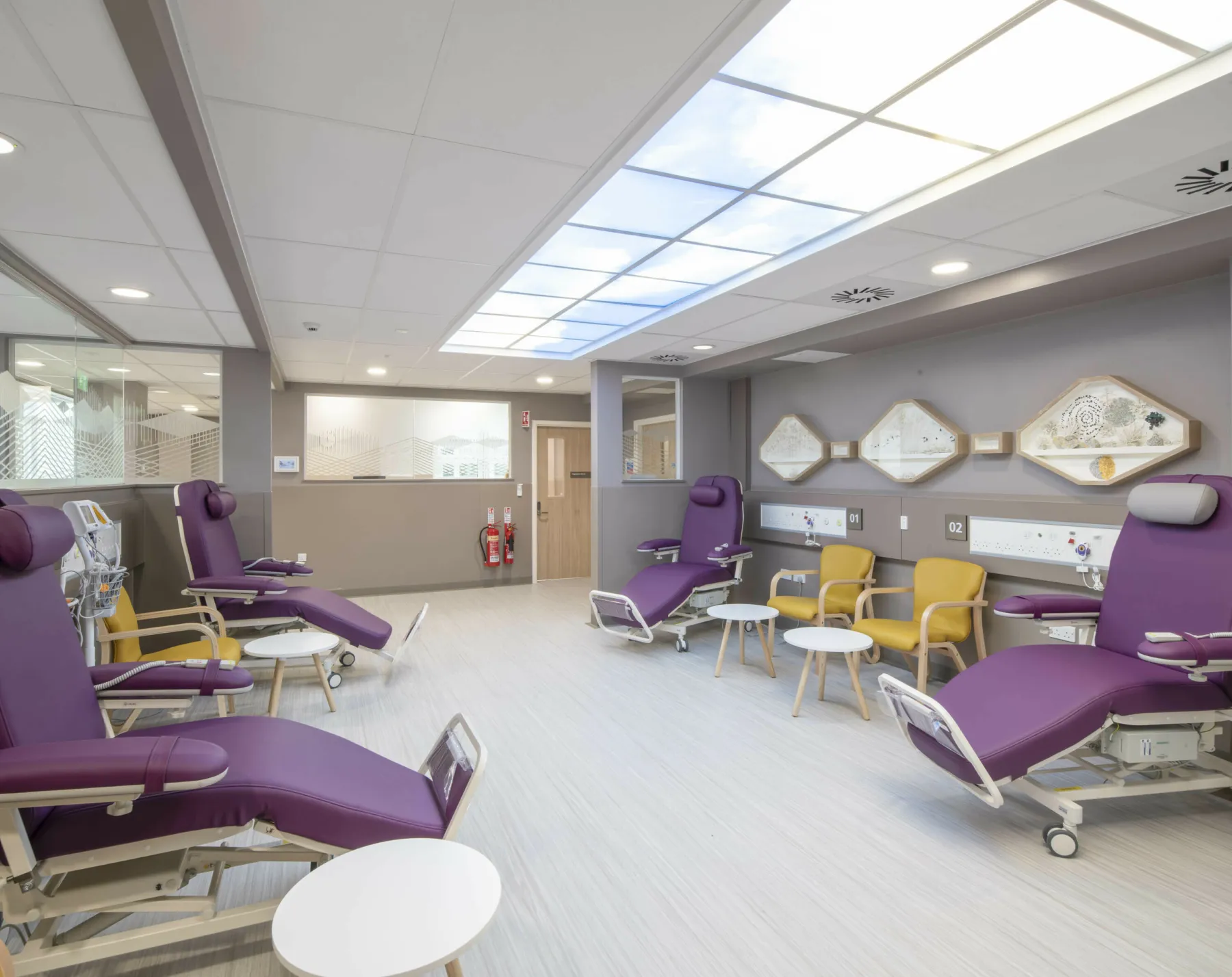 A treatment ward in the Haematology Centre at the Western General Hospital, Edinburgh. Chairs for patients and visitors are arranged around the room. Bespoke artwork is on the walls and a bespoke installation provides the illusion of a skylight above.