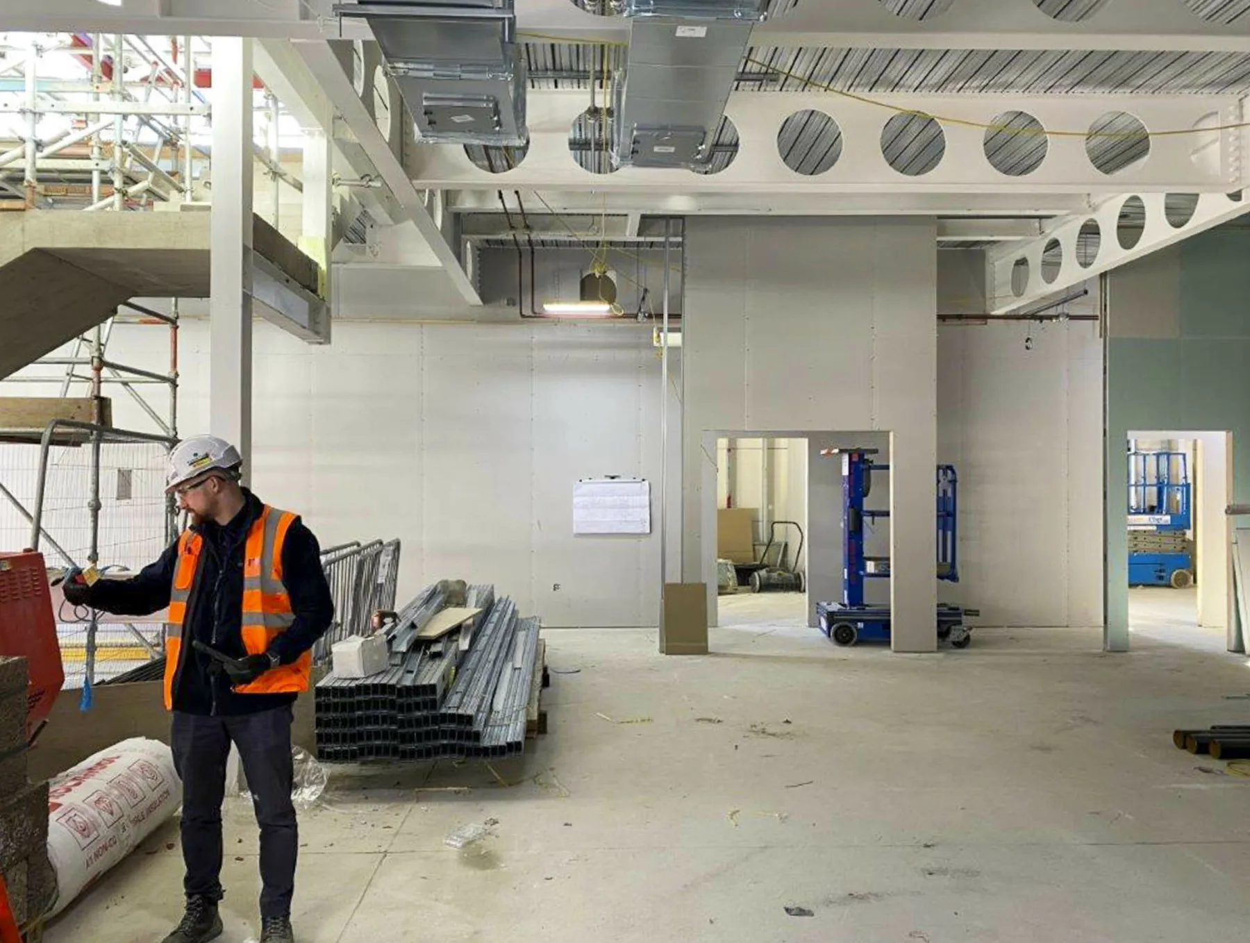 On a construction site at the Western General Hospital Edinburgh. Inside a building a male worker wears an orange hi vis vest and white hard hat. Materials are waiting to be used and the building is still in shell form.
