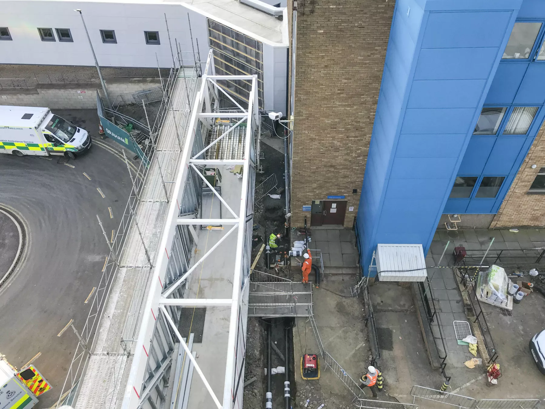 Aerial view showing bridge link being put in place at the National Treatment Centre - Fife Orthopaedics. Behind the link, contstruction workers are wearing orange hi vis clothing. On the other side, ambulances are parked.