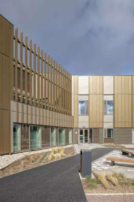 Exterior view of the National Treatment Centre - Highalnd, Inverness. Gold-bronze coloured cladding with a perforated screen for the first floor courtyard garden. Paths and landscaped beds with grasses.