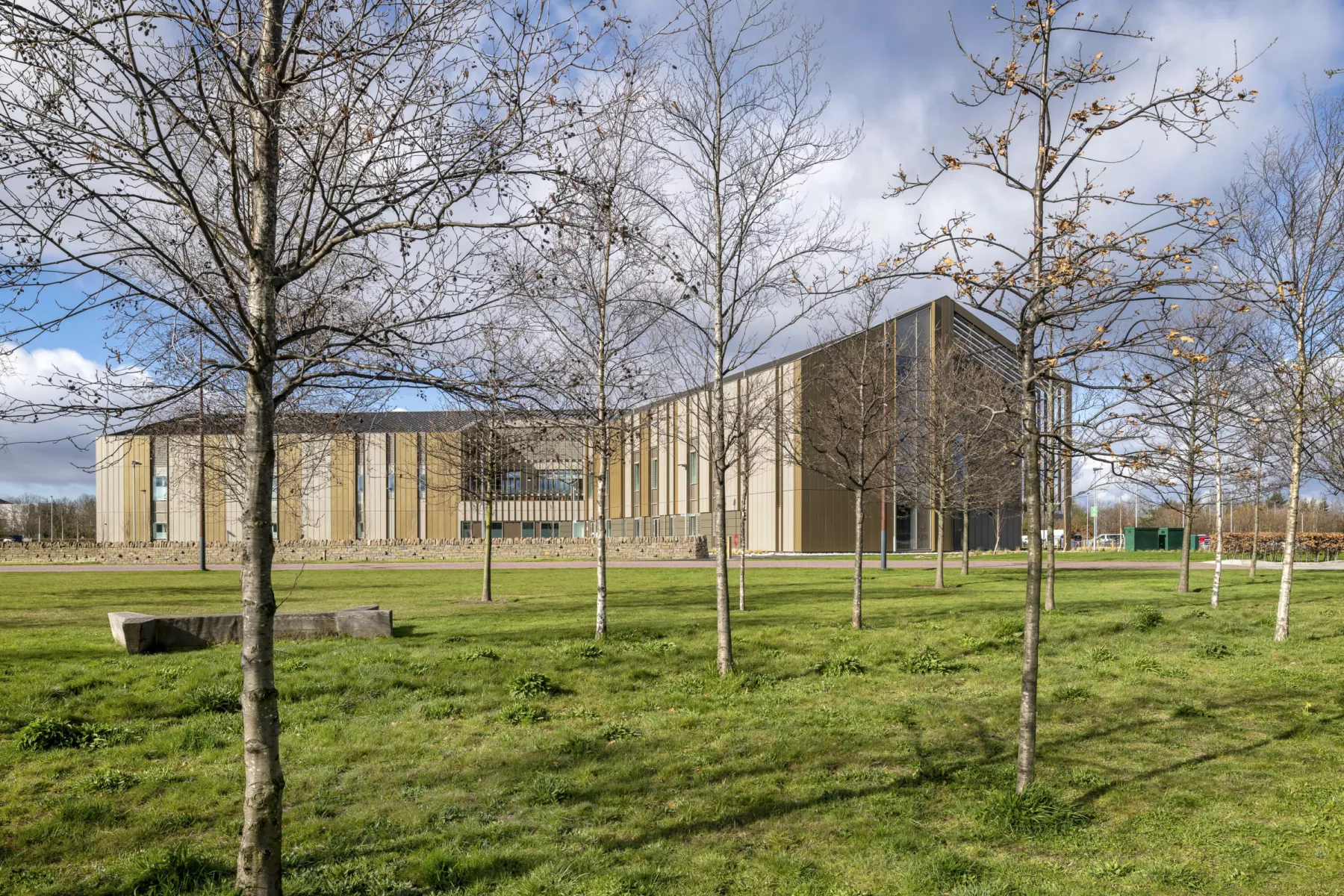 National Treatment Centre - Highland, a new hospital building in Inverness. See through landscaped area planted with trees, the building is clad in a golden coloured metal and has a pitched zoof.