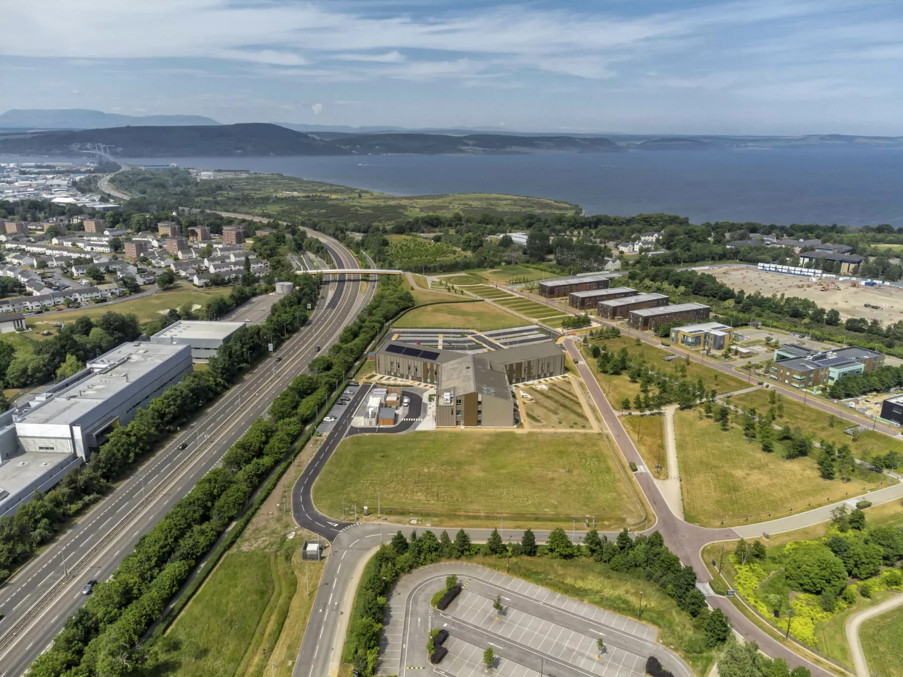 Aerial view of the National Treatment Centre - Highland, Inverness. Showing the new healthcare centre surrounded by green space and with a dual carriageway to the left hand side of the image. In the distance the sea.