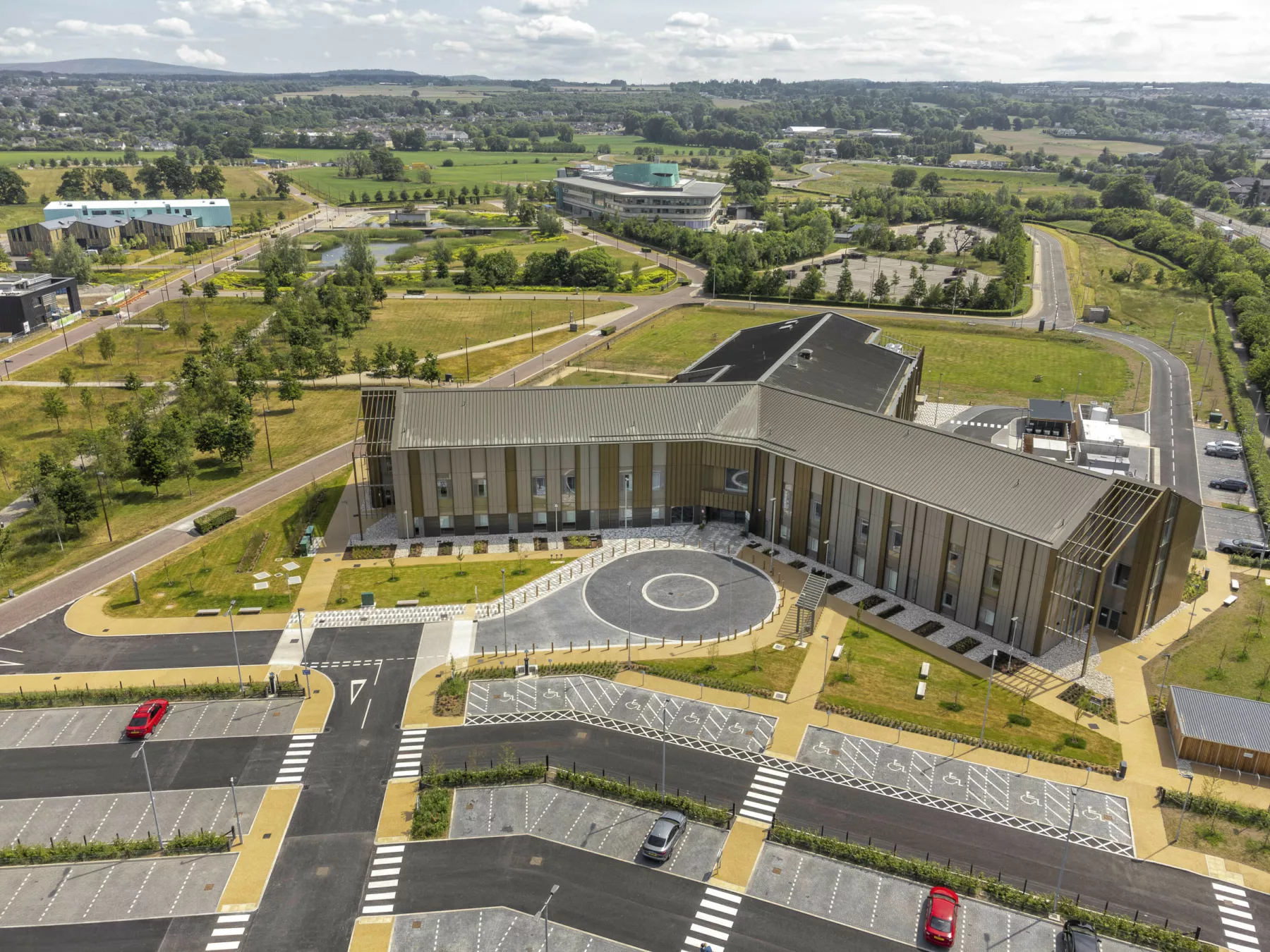 Aerial view of the National Treatment Centre - Highland, Inverness. The building's three wings form a triad in landscaped grounds. The building has a pitched roof and is clad in gold-bronze metal cladding. Car parks and surround lightly wooded areas are shown, with other buildings on the campus in the distance