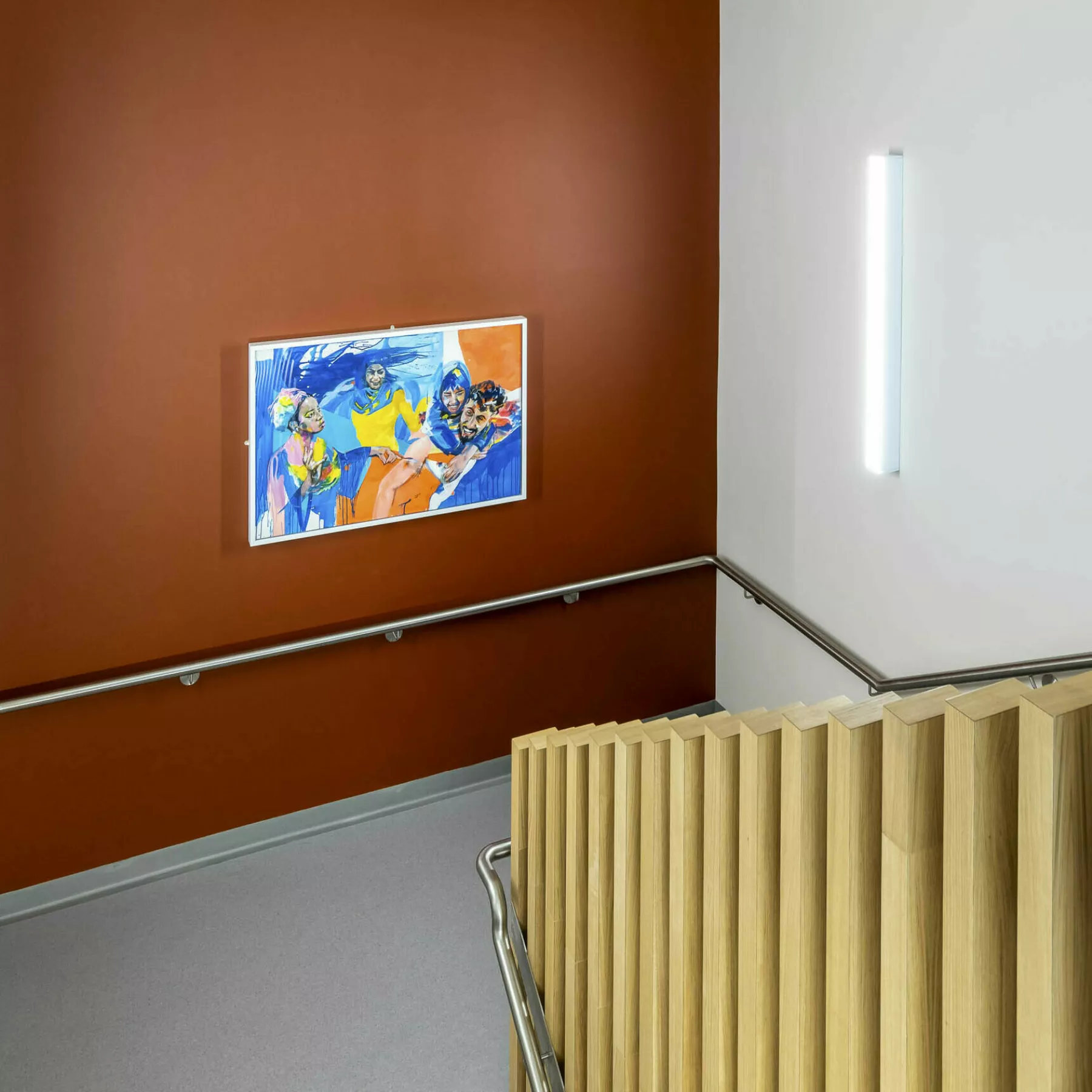 View of stairwell at the National Treatment Centre - Highland. A dark orange wall has a bright, contempoary painting. The bannister and ballustrade is wood with a steel rail.