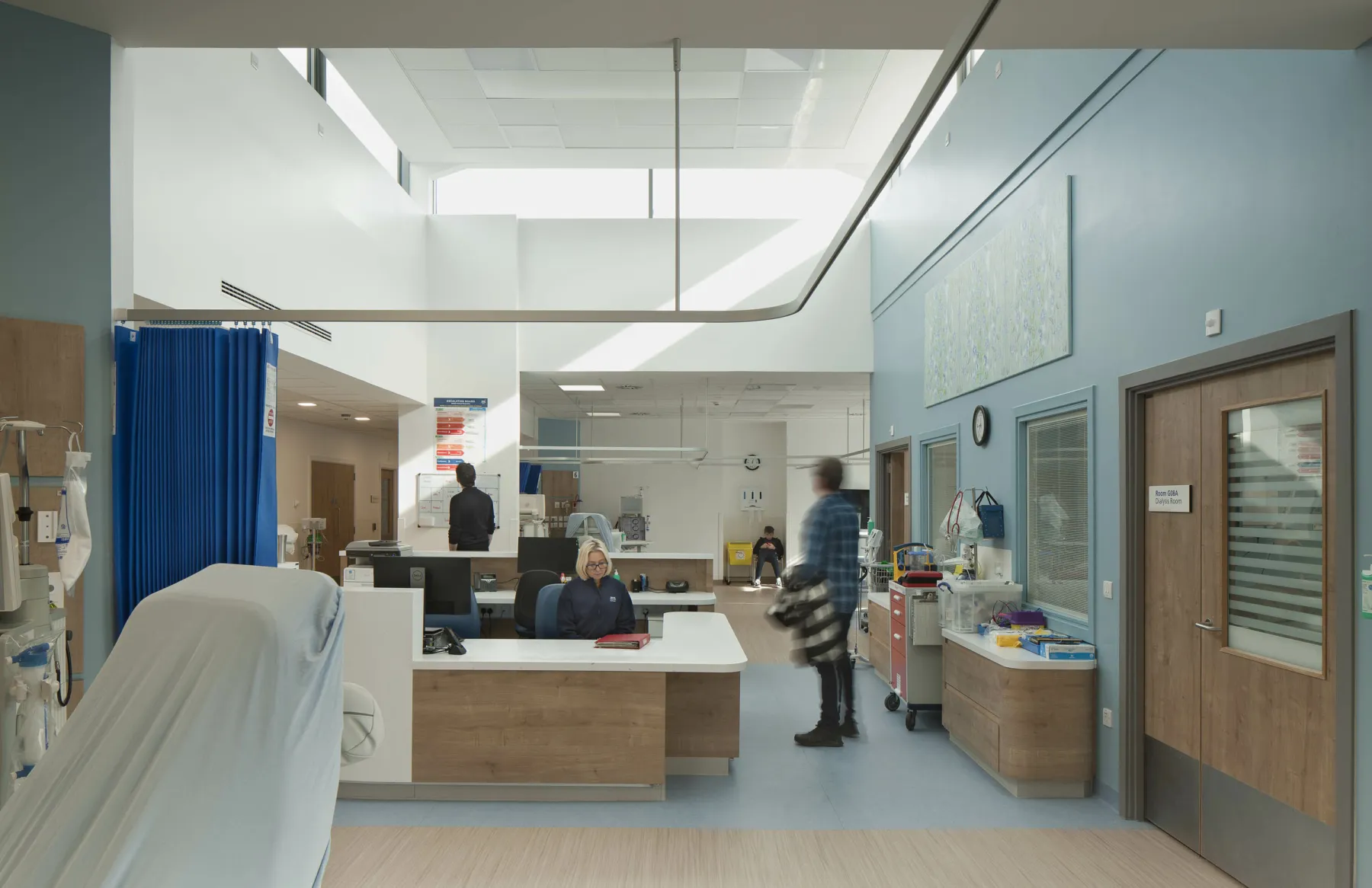 At reception in the Renal Dialysis Unit, Edinburgh - a purpose built healthcare facility at the Western General Hospital. A member of staff receives a visitor at the reception desk in the double height, naturally lit space.