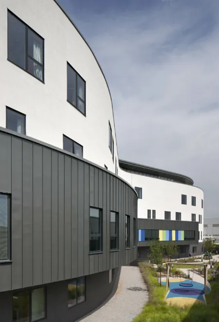 Detail of the curved exterior of the Royal Hospital for Sick Children, Edinburgh, newly finished. The lower storeys are clad in dark grey, with upper storeys in white render. Window frames are dark grey. A garden and play area is shown on the right.