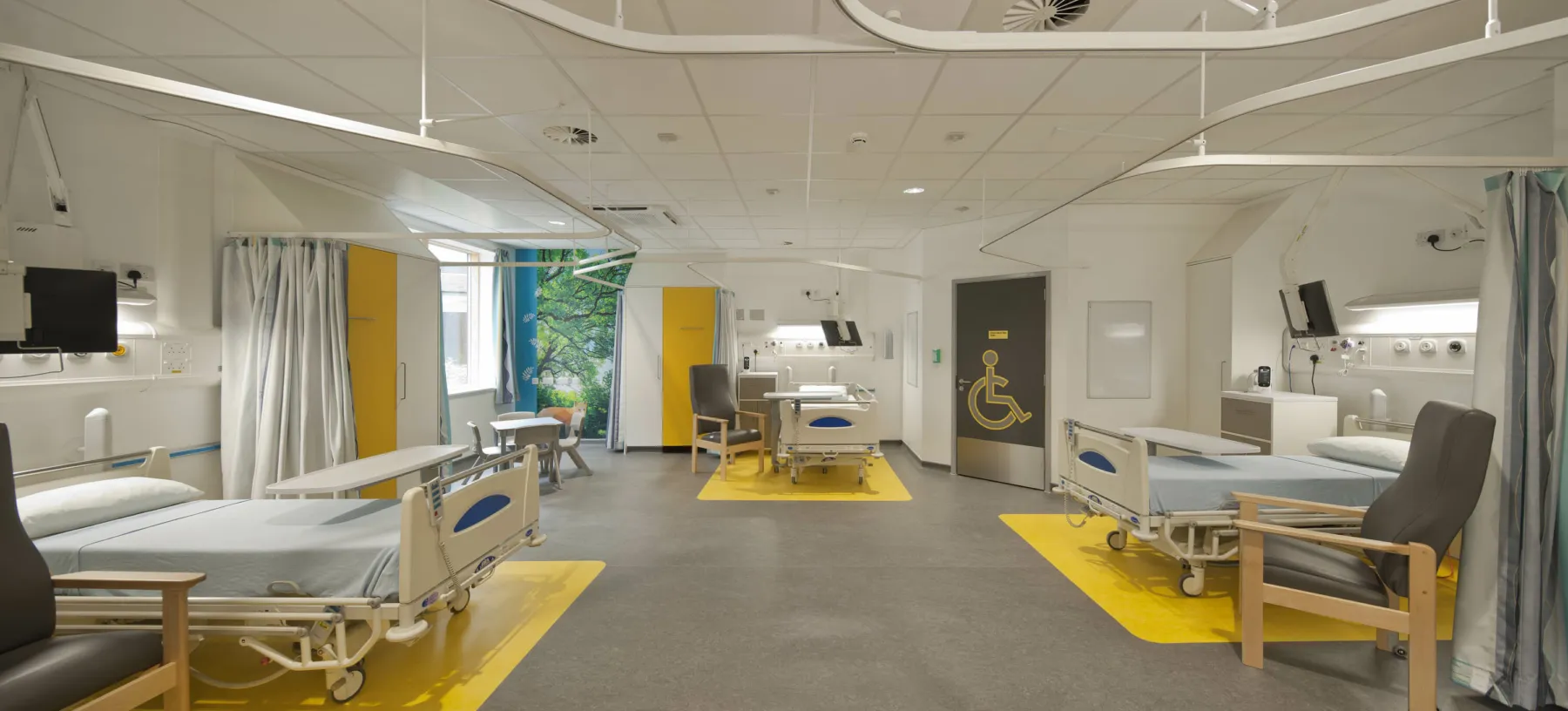 In one of the wards at the Royal Hospital for Sick Children, Edinburgh.  Three beds are arranged with space around them and chairs for visitors. The floor is coloured yellow underneath the beds and some wall murals add interest to the space.