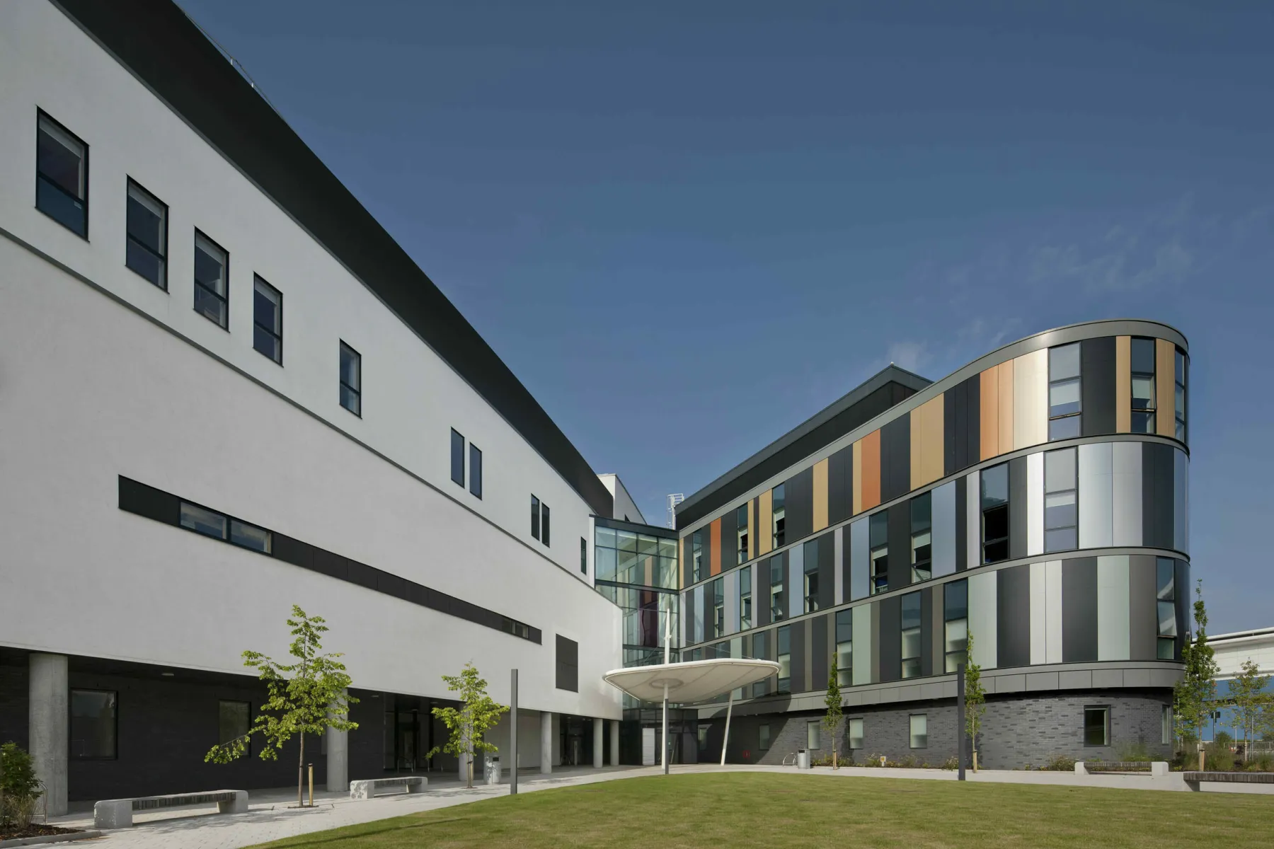 View of the exterior of the Royal Hospital for Sick Children And Young People, Edinburgh. Landscaped areas and trees sit along side two sections of the building  which meet at a canopied entrance. On the left the building has white render, on the right a bow-ended building is clad in copper and silver tones with large windows.