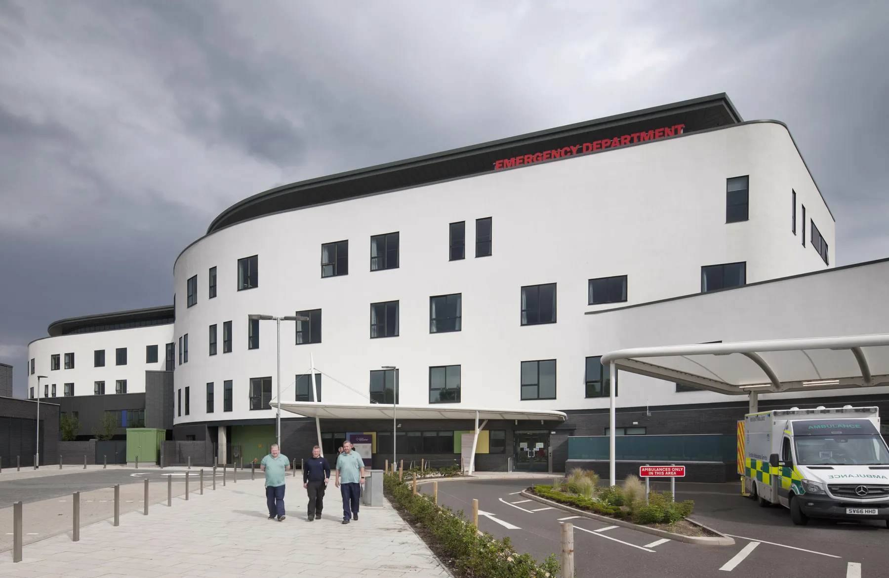 The exterior view of the Emergency Department at the Royal Hospital for Sick Children and Young People, Edinburgh. A White building with 5 storeys, curved ends and irregularly placed windows. There is a canopy above the door for admissions. Three staff members walk towards the camera.