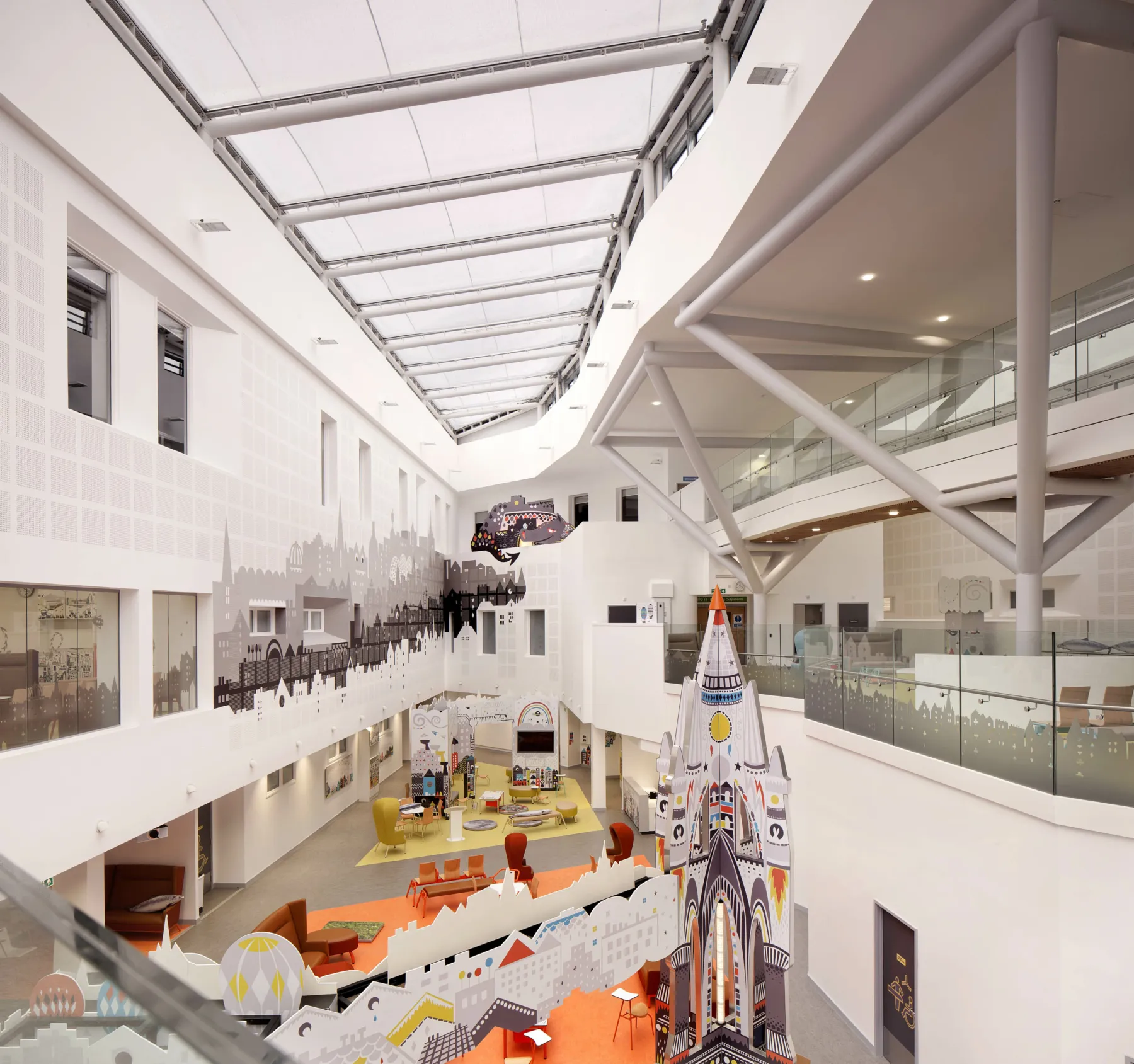 A view into the atrium at the Royal Hospital for Sick Children, Edinburgh, from the first floor gallery. A glass roof creates a brightly lit space with sculpture, a wall mural colour and playful design features.