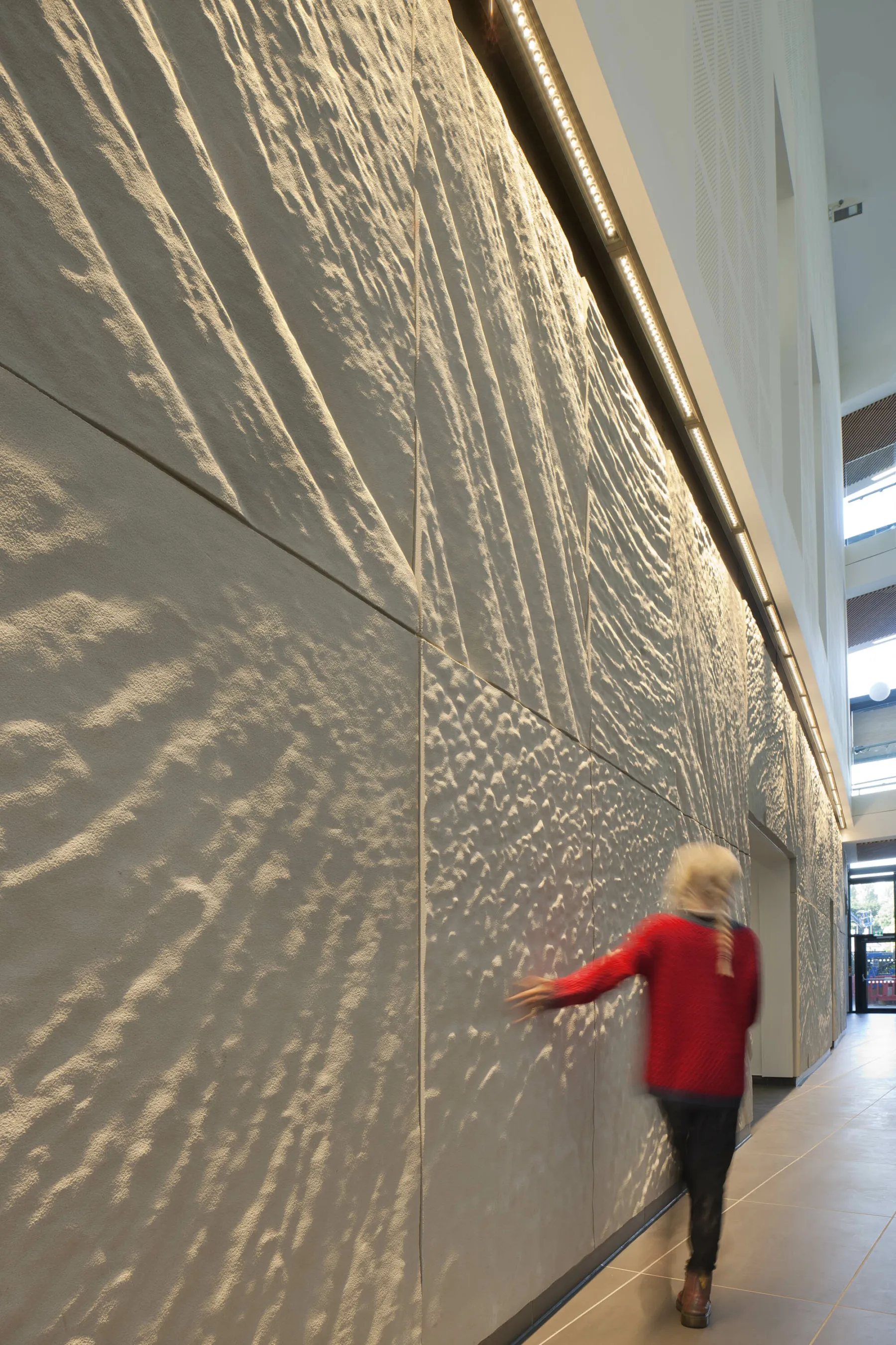 A wall is clad in a tactile sandstone-like surface - which has the appearance of a fossilised sea bed. A young girl walks past running her hand over it.