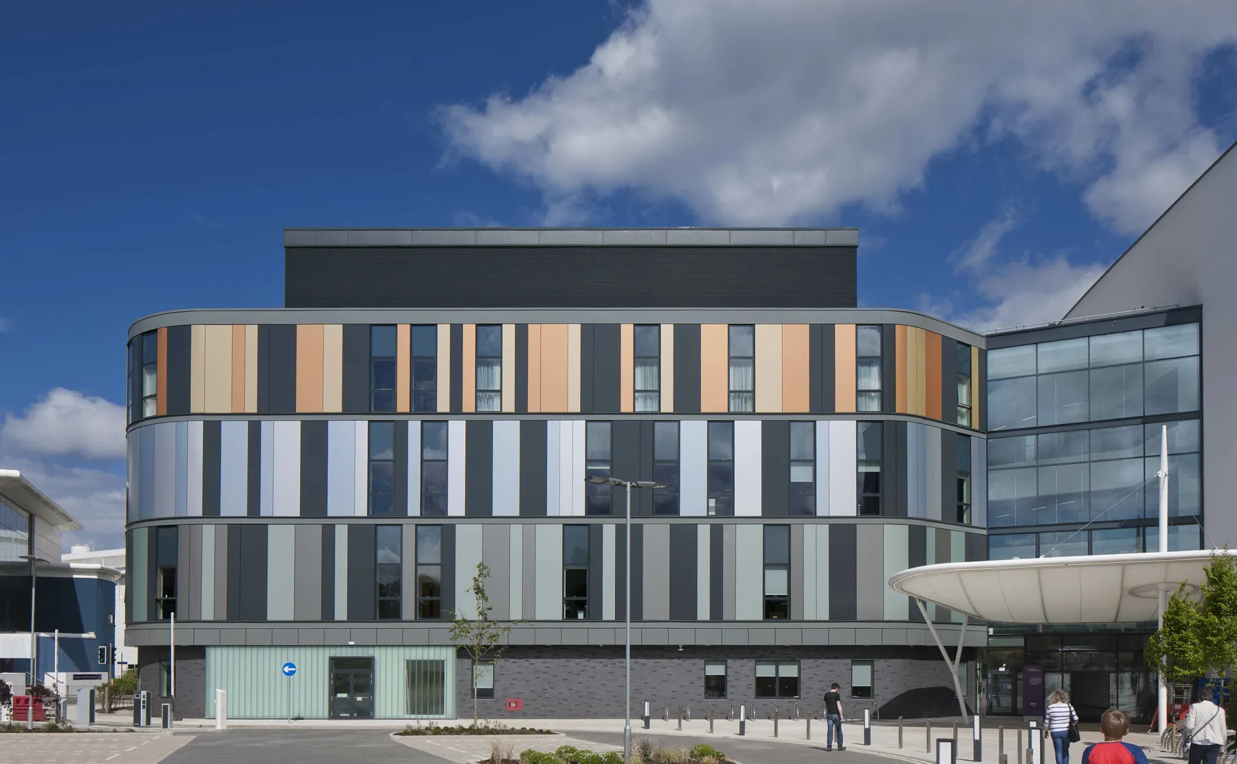 A bow-ended building at the Royal Hospital for Sick Children and Young People, Edinburgh. A purpose build new hospital, the main entrance with canopy is shown. The four storey building in the centre of the image is clad in copper, grey and green tones.