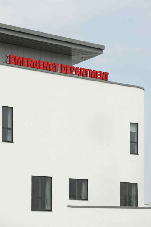 Exterior detail of the Emergency Department of the Royal Hospital for Sick Children and Young People, Edinburgh. A large, illuminated red sign "Emergency Department" sits above the main entrance to the white-rendered building.