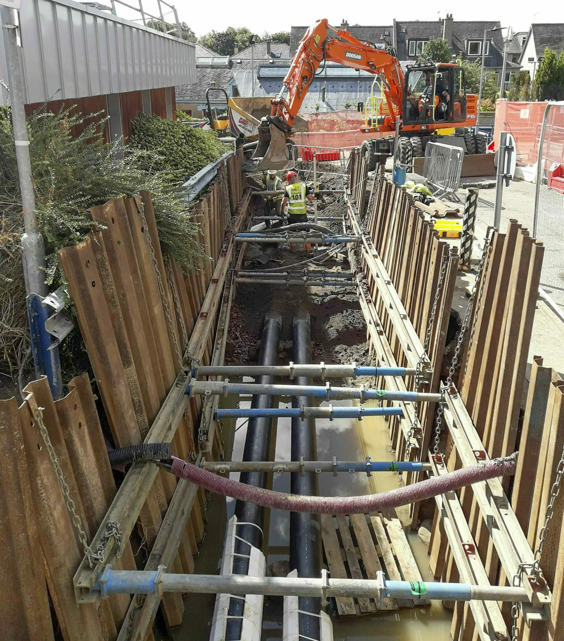 Energy Infrastructure upgrades at the Western General Hospital Edinburgh. A deep trench with reinforced edges. Inside various sections of piping are being installed. A digger is in the background.