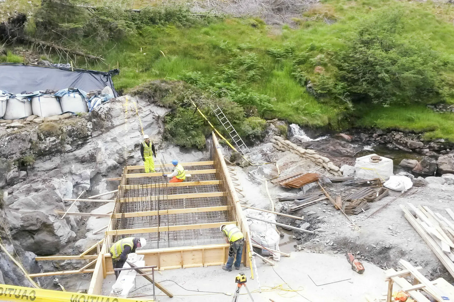 A hydro scheme under construction in Scotland. A timber frame is built across a river supported by concrete. Four workers in hi-vis are position across the structure. In the background rock that has been cut into  is visible.