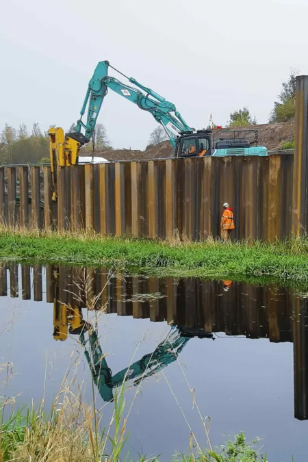 Diggers at work behind a temporary steel retaining wall at the forth and clyde canal, Glasgow. Construction site for the Stockingfield Bridge. A construction worker walks past wearing orange hi-vis clothing.