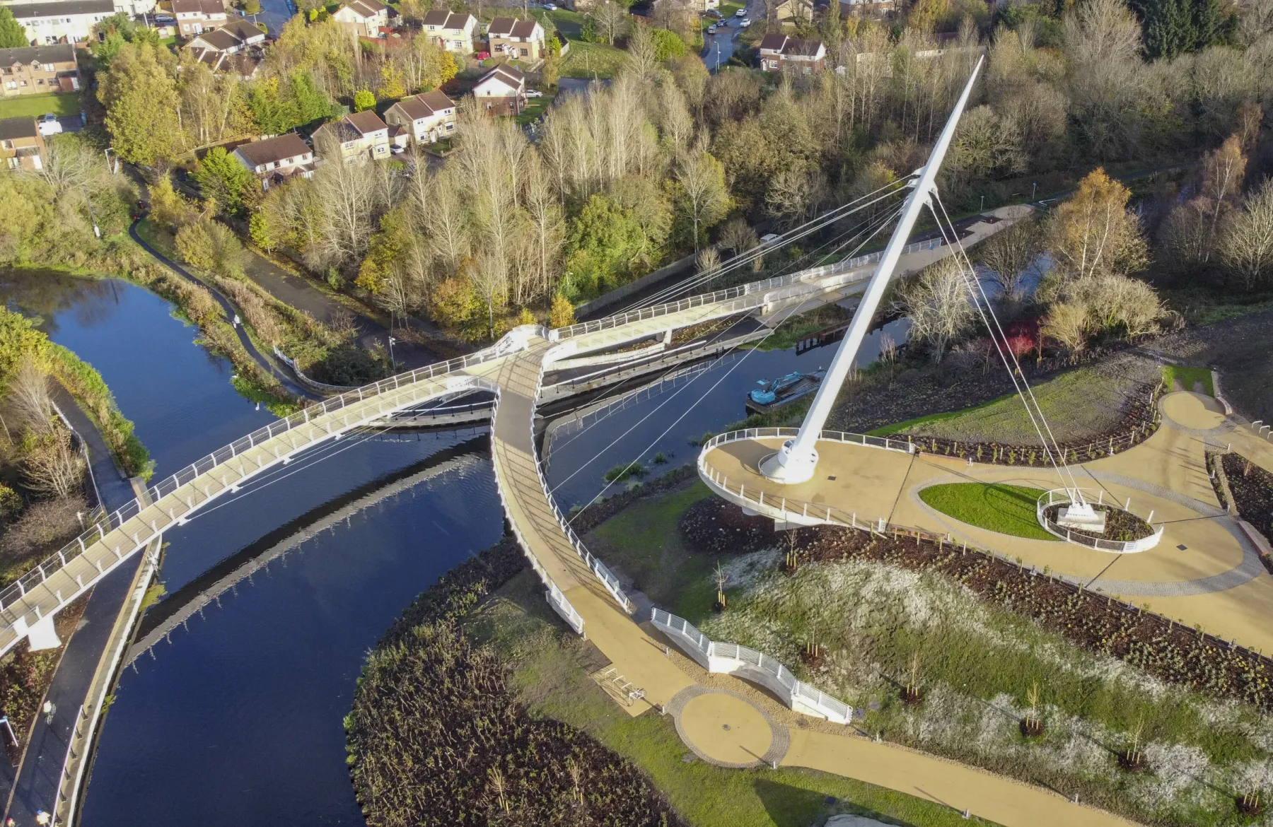 An aerial view of the Stockingfield Bridge, Glasgow. The cable-stayed pedestrian and cyclist footbridge connects three areas across a forked junction in the forth-clyde canal. The decks of the bridge are shown stretching across the canal, and landscaping around the tower and bridge entrances.