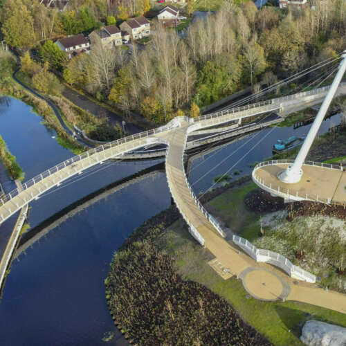 An aerial view of the Stockingfield Bridge, Glasgow. The cable-stayed pedestrian and cyclist footbridge connects three areas across a forked junction in the forth-clyde canal. The decks of the bridge are shown stretching across the canal, and landscaping around the tower and bridge entrances.