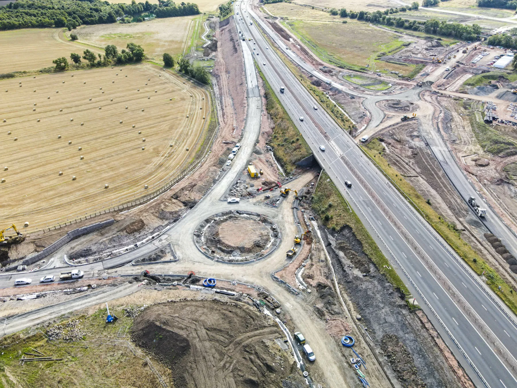 An aerial view of the M9 junction at Winchburgh under construction. The motorway flies over a road with two feeder roundabouts. The road works are surrounded by fields at harvest with hay bales.